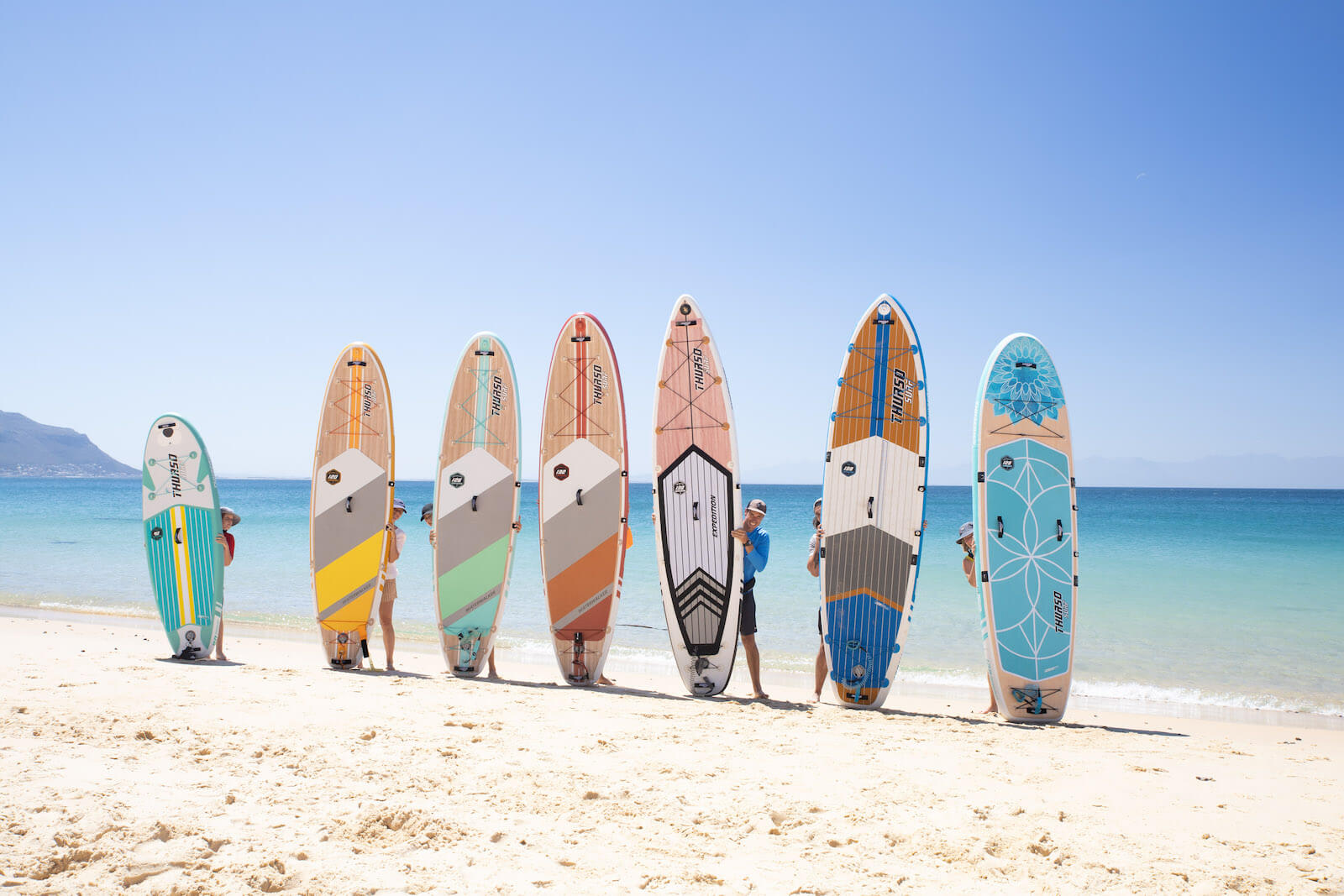 7 Thurso Surf stand up paddle boards being held by paddleboarders on the beach